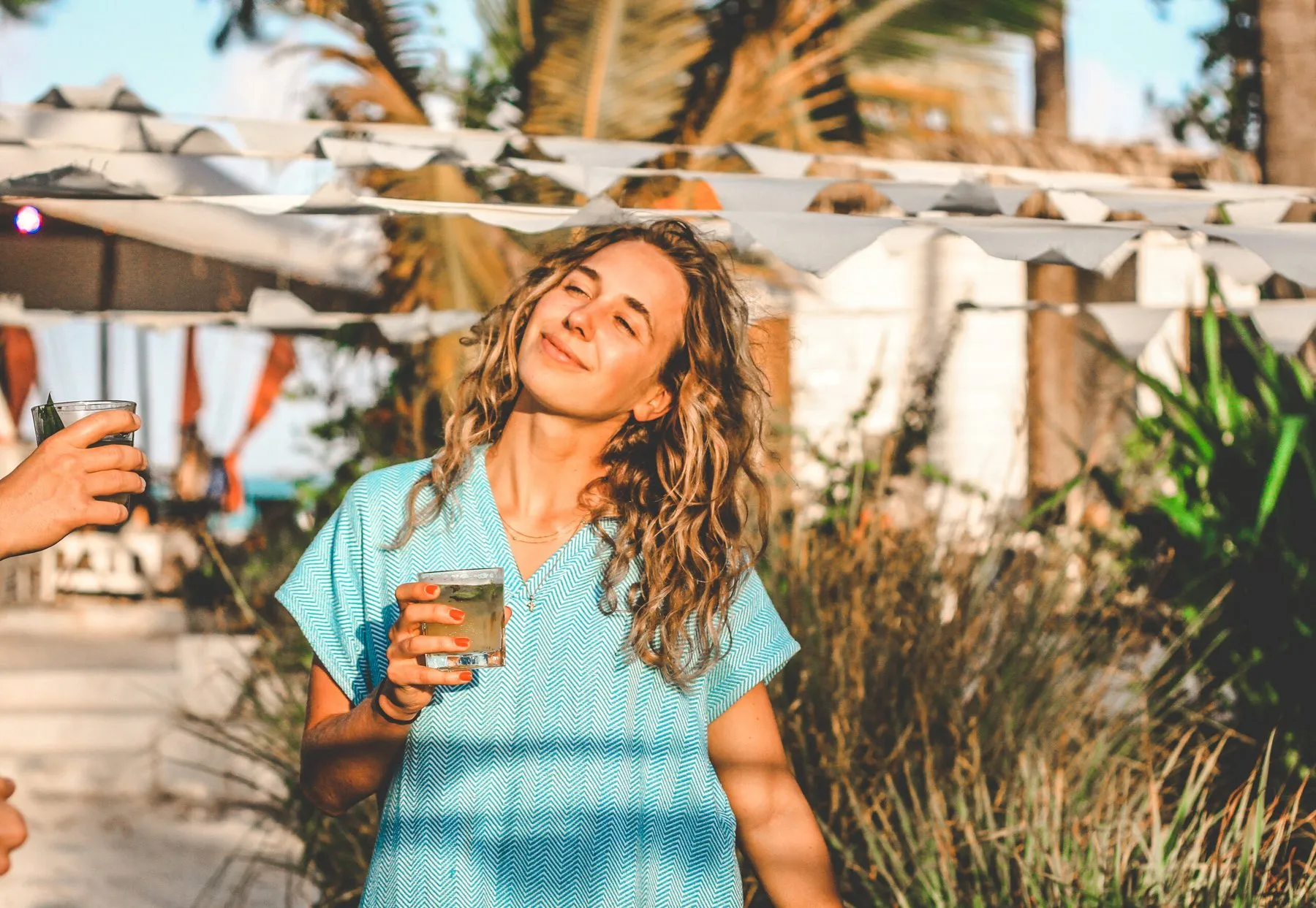 Junge Frau mit Locken und Getränk in Strandbar bei Sonnenuntergang