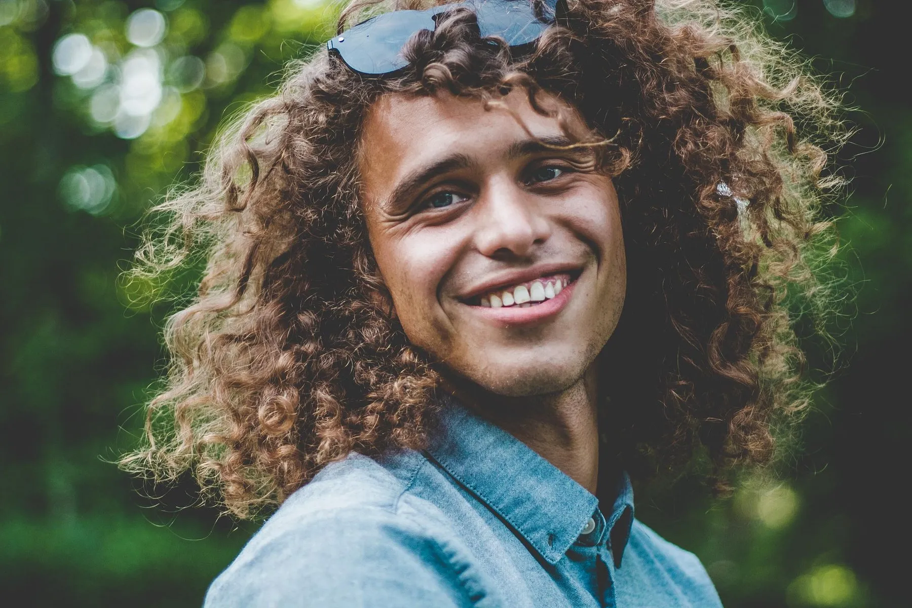 Lachender junger Mann mit Locken und Sonnenbrille im Grünen