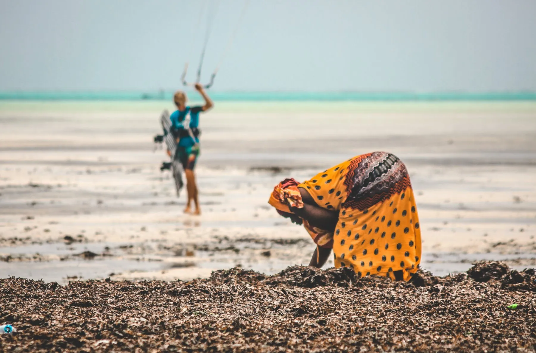 Frau in buntem Kleid sammelt Algen am Strand, Kitesurfer im Hintergrund