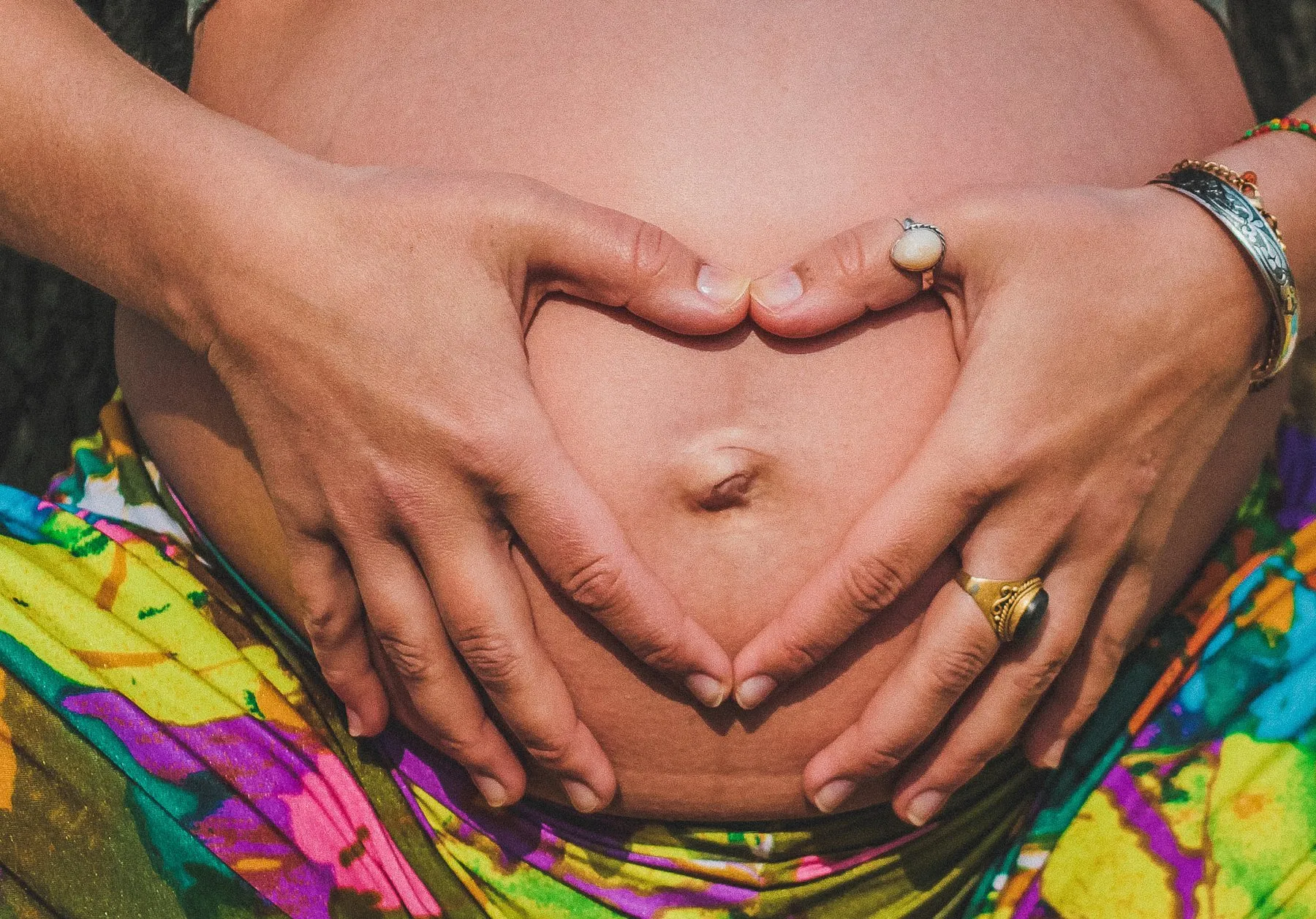Schwangerer Bauch mit Händen in Herzform vor buntem Wickeltuch